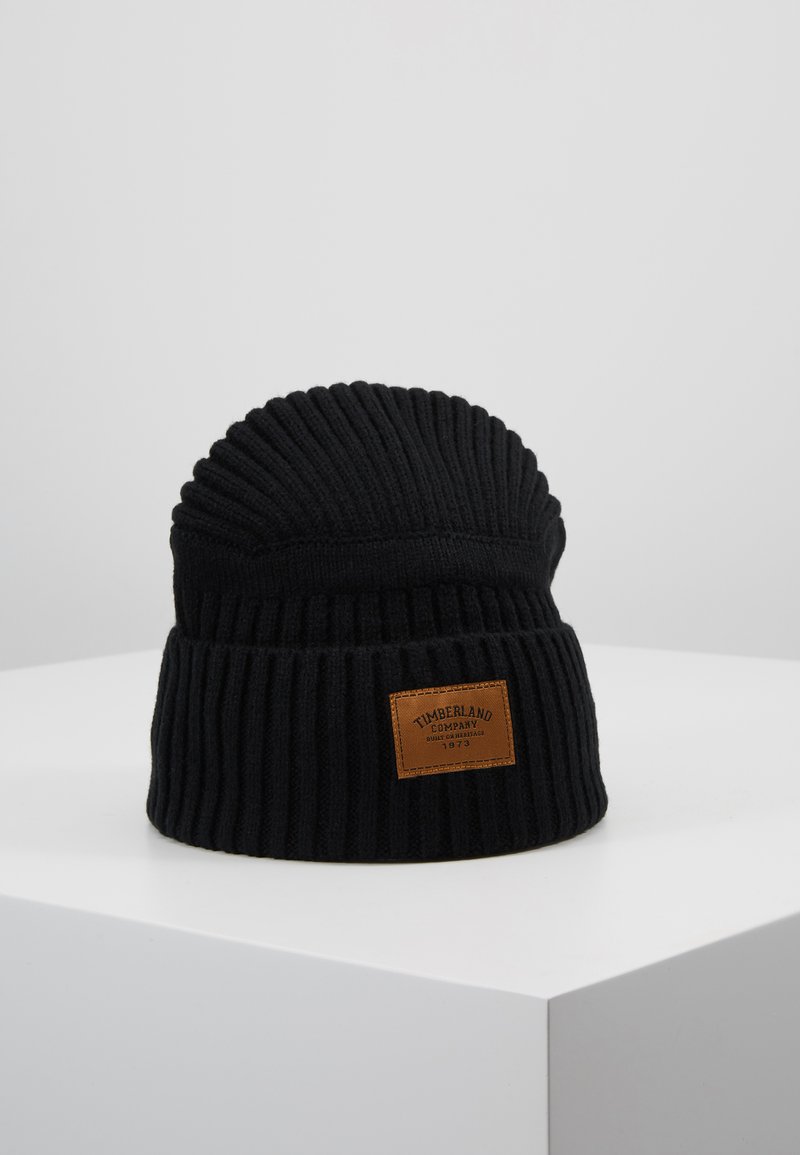 Mustat ribbed-kudotut pipo, jossa on taitettu huppu ja suorakulmainen ruskea nahkalappu, jossa on "Timberland Company" -logo.