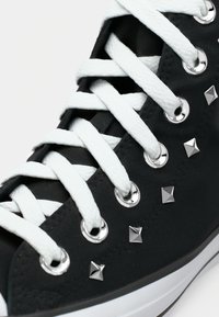 Adidași negri din pânză cu glezna înaltă, cu șireturi albe, ochiuri argintii și ținte piramidale pe laterale. Talpă din cauciuc alb. Vedere detaliată.