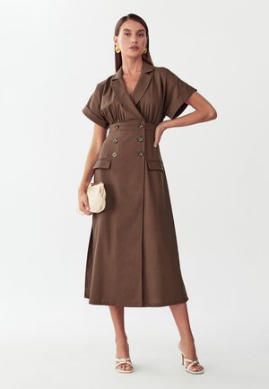 Frau trägt ein braunes, zweireihiges Midikleid mit kurzen Ärmeln, hält eine beige Clutch und trägt weiße Sandaletten mit Absatz und goldene Ohrringe.