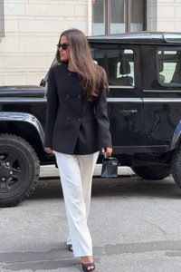 Svart dubbelknäppt kavaj, vita skräddarsydda byxor, svarta sandaler med öppen tå, håller en liten svart handväska, med en SUV i bakgrunden.