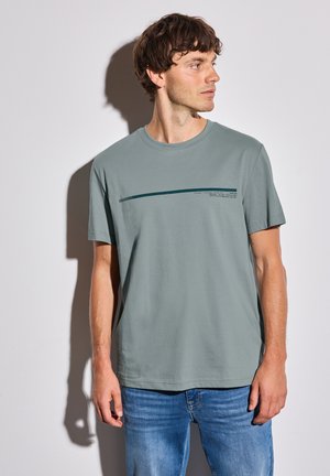 T-shirt en coton vert clair avec un col rond, présentant un imprimé graphique horizontal vert foncé sur la poitrine, associé à un jean bleu.