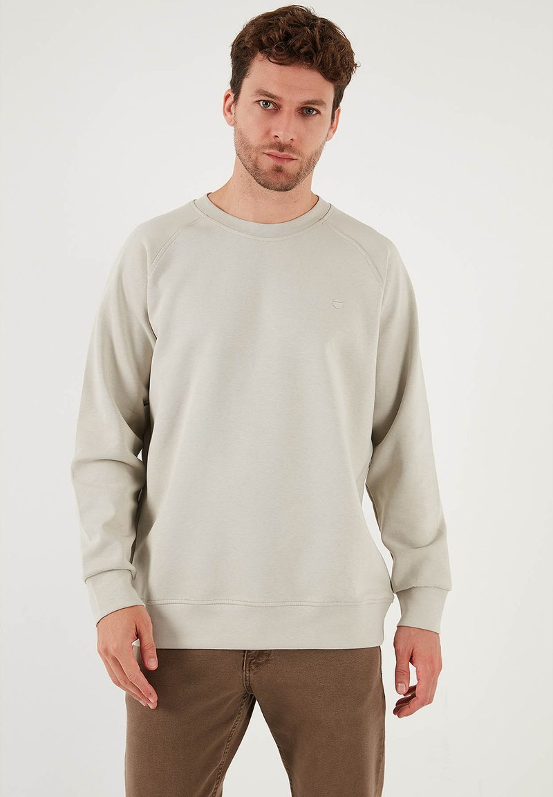 Felpa leggera grigia a girocollo, con maniche raglan, texture morbida, minimal branding sul petto, abbinata a pantaloni marroni.