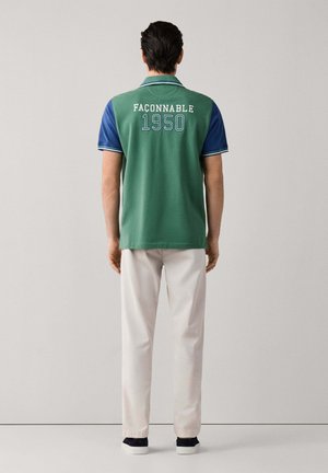 Man standing facing away wearing green and blue polo shirt with "Faconnable 1950" text on back, white pants, and dark shoes.