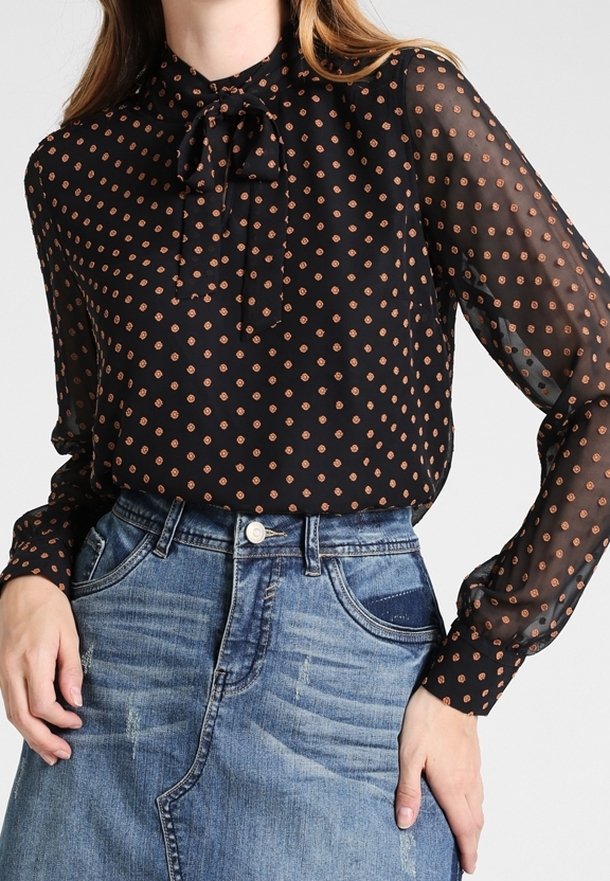 Woman wearing sheer black blouse with orange polka dots and a self-tie bow neck, paired with a high-waisted blue denim skirt.