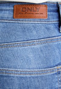Šviesiai mėlyni džinsai turi odinį ženkliuką su užrašu "ONLY" ir "Blue Denim since 1995." Aiškiai matomi siūlai aplink liemenį.