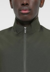 Κοντινό πλάνο ενός ατόμου που φοράει ένα ανοιχτό πράσινο μπουφάν με κλειστό φερμουάρ και ψηλό γιακά, δείχνοντας το κάτω μέρος του προσώπου και τον λαιμό.