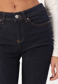 Jeans de mezclilla azul oscuro con costuras naranjas, cierre frontal de botón y bolsillos estándar, con un corte recto y una textura suave.