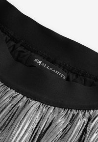 Ceinture élastique noire avec une texture froncée sur le dessus, menant à un matériau à franges métalliques argentées. Étiquette de marque visible.