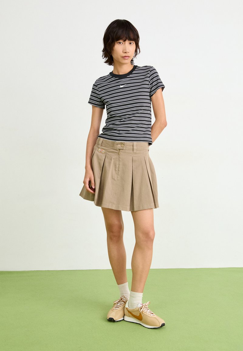 T-shirt rayé noir et blanc, jupe plissée beige, baskets beiges avec accents orange, situées sur un sol vert.