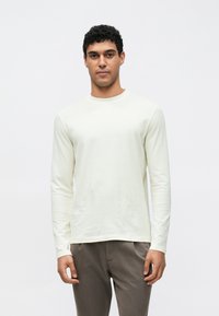 Hellcremefarbenes langärmliges Baumwollshirt mit rundem Ausschnitt und glatter Textur, kombiniert mit grauen, maßgeschneiderten Hosen.