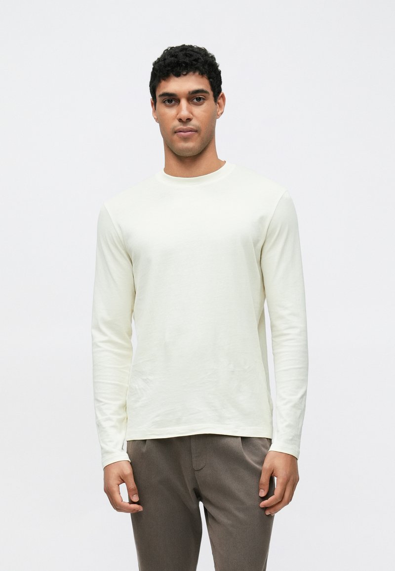 Hellcremefarbenes langärmliges Baumwollshirt mit rundem Ausschnitt und glatter Textur, kombiniert mit grauen, maßgeschneiderten Hosen.