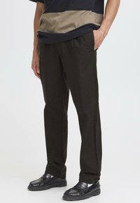 Una persona está de pie vistiendo pantalones oscuros y una camisa a rayas de colores con secciones marrones y negras, combinada con zapatos negros.