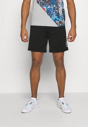 Männlich, trägt schwarze Sportshorts mit weißem Text an der Seite, ein graues und mehrfarbiges abstraktes Oberteil und weiße Sportschuhe mit Schnürsenkeln.