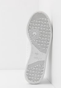 Suola di sneaker bianca con motivo a trama circolare, superficie testurizzata e un logo minimale. Accentuata da leggeri spruzzi e linee pulite.