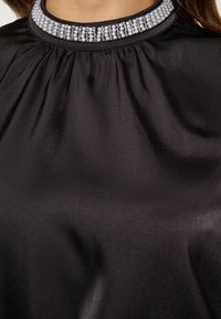 Top in satin nero con scollatura arricciata, caratterizzato da un pannello decorativo con strass incorporati attorno al colletto. Texture liscia.