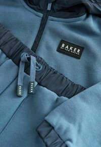 Blauwe hoodie met een gestructureerde stof en een aangesloten ontwerp. Voorzien van een zwart label met "BAKER" en een strakke trekkoord bij de kraag.