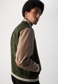 Chaqueta de béisbol de mezcla de lana verde con mangas de cuero marrón, puños acanalados y cuello a rayas. Presenta un detalle bordado en la parte posterior.