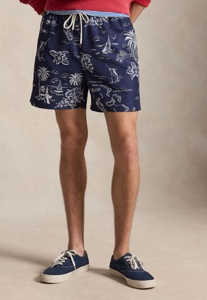 Άνδρας που φοράει σορτς με τροπικό μοτίβο σε ναυτικό μπλε χρώμα και ναυτικό μπλε αθλητικά παπούτσια με λευκά κορδόνια, στέκεται μπροστά σε μονόχρωμο μπεζ φόντο.