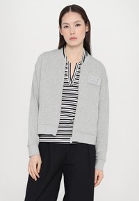 Šedá zkrácená bomber bunda z měkké tkaniny, s oboustranným zipem a žebrovanými manžetami, nošená přes top s černobílými pruhy.