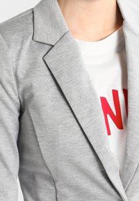 Blazer gris clair à revers crantés et tissu texturé, porté sur un T-shirt blanc avec un texte rouge.