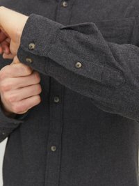 Camisa de franela gris oscuro con puños abotonados y bolsillo en el pecho. Presenta una tela texturizada y un collar redondeado con botones a juego.