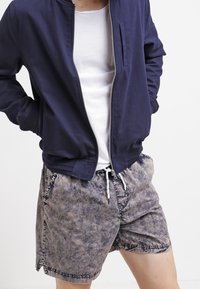 Veste de bombardier navy avec une fermeture éclair sur le devant, des poignets côtelés et une poche sur la poitrine, accompagnée de shorts en toile à cordons, de couleur brun clair et délavés.