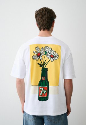 Bărbat purtând tricou alb cu grafică cu sticla de 7up ținând trei margarete albe pe fundal galben.
