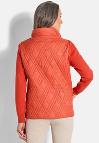 Frau trägt eine leuchtend orange gesteppte Weste mit Rautenmuster über einem Langarmshirt, steht mit der rechten Hand auf der Hüfte, mit dem Rücken zum Betrachter.
