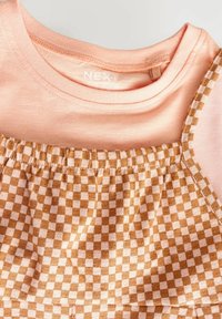T-shirt en coton rose superposé sous un tissu à carreaux marron et crème avec un effet froncé élastique, présentant des bretelles fines pour un look superposé.