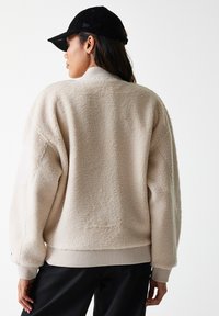 Beige fleece-takki rentoa istuvuutta, ribbaustakkeja ja helmaa sekä pehmeää rakennetta. Yhdistetty mustan lippiksen kanssa, joka esittelee minimalistista muotoilua.