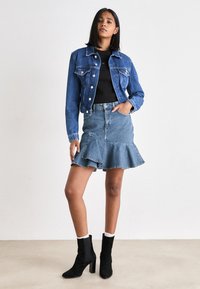 Denimist jakistusega jope, mis on kinni nööbiga, koos denimsärgiga, millel on laienev äär. Mustad pahkluu saapad tekstureeritud viimistlusega ja valged sokid.