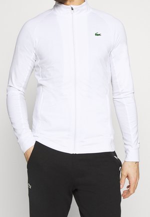 Homme portant une veste blanche zippée avec un logo crocodile vert sur la poitrine et un pantalon noir, les mains dans les poches.