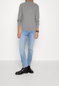 Grauer gerippter Pullover mit einem halben Reißverschlusskragen, kombiniert mit hellblauen geraden Jeans und schwarzen Leder-Stiefeletten.