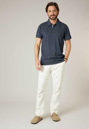 Man met baard die een donkerblauw poloshirt met korte mouwen, witte broek en beige instapschoenen draagt, staand met één hand in de zak tegen een effen achtergrond.