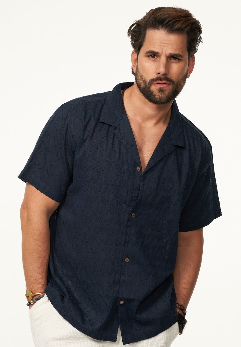 Marineblaues Kurzarm-Hemd mit Knöpfen und strukturiertem Blumenmuster, lässiger Schnitt und braunen Knöpfen. Getragen mit weißen Hosen und Accessoires.