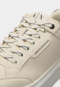 Αθλητικό παπούτσι από κρεμ δέρμα με κορδόνια σε μαύρο και λευκό, με ανάγλυφο το "Armani Exchange" στο πλάι και λογότυπο "A|X" στη γλώσσα.