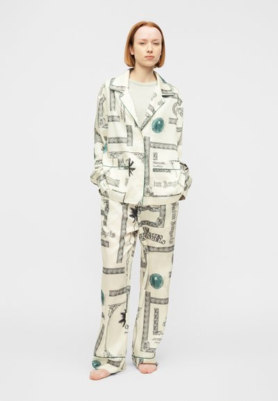 Νεαρό άτομο που στέκεται ξυπόλυτο, φοράει ένα φαρδύ λευκό κοστούμι με μαύρα και τυρκουάζ γραφικά σχέδια και ανοιχτό γκρι πουκάμισο από κάτω.