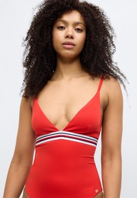 Κόκκινο ολόσωμο μαγιό με βαθιά V-λαιμόκοψη και λεπτές τιράντες. Διαθέτει οριζόντια ριγέ διακοσμητικό σε μαύρο και άσπρο γύρω από τη μέση.