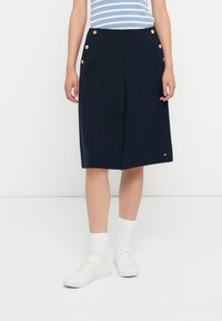 Námořnická midi sukňa vyrobená z textúrovanej tkaniny, s predným sklonom a zlatými gombíkovými akcentmi na páse. Nosená s bielymi teniskami a ponožkami.