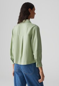 Hellgrüne Bluse mit langen Ärmeln und lockerer Passform, Knopfmanschetten und einem gepleckten Rücken. Kombiniert mit blauen Jeans, was einen lässigen Look zeigt.