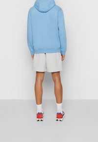 Ljust blå bomullshuvtröja med dragsko på huvan, kombinerad med grå shorts och röda sportskor. Synlig textur och avslappnad passform.