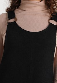 Czarna, teksturowana sukienka ogrodniczka z regulowanymi ramiączkami i złotymi elementami, noszona na różowej, prążkowanej bluzce z golfem.