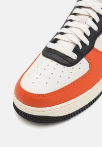 Zapatilla blanca con acentos en naranja y negro, con una parte superior de cuero texturizado, puntera perforada y suela de goma negra.