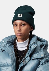 Gorro verde oscuro con textura acanalada y parche de logo blanco, combinado con una chaqueta acolchada azul claro y un suéter negro debajo.