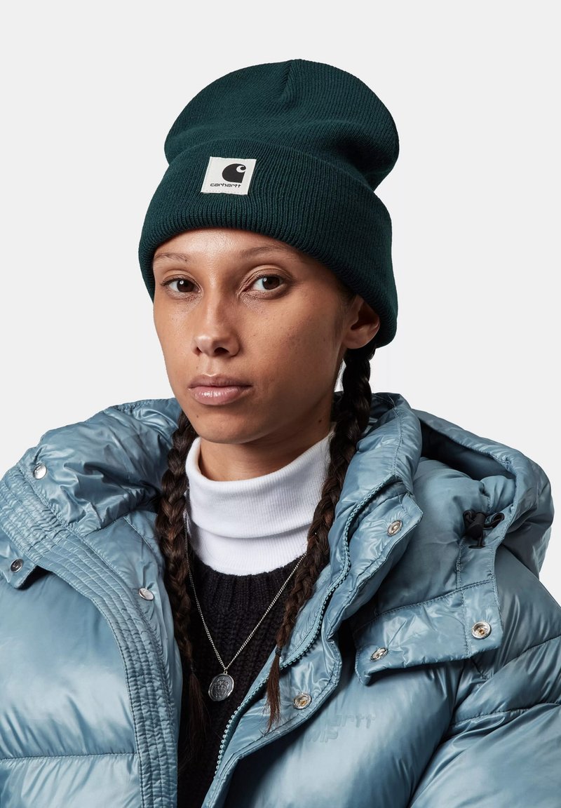 Gorro verde oscuro con textura acanalada y parche de logo blanco, combinado con una chaqueta acolchada azul claro y un suéter negro debajo.