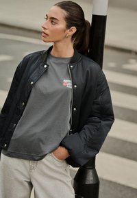 Sötét tengerészkék bomber dzseki cipzárral, ezüst színű fém részletekkel és fekete kiemelésekkel, rajta egy szürke póló virágos szöveggel. Világosszürke farmer