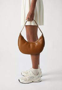 Sac en cuir marron avec une forme incurvée et une texture lisse, tenu par une personne portant une robe blanche et des baskets claires.