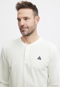 Hvit langermet henley-skjorte laget av myk stoff, med en tre-knapps placket og et trekantet logo-patch på brystet.