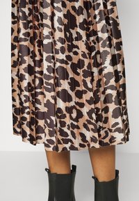 Gonna midi plissettata in tessuto morbido con sfondo beige e stampa leopardo marrone scuro. Stivali neri alla caviglia visibili in basso.