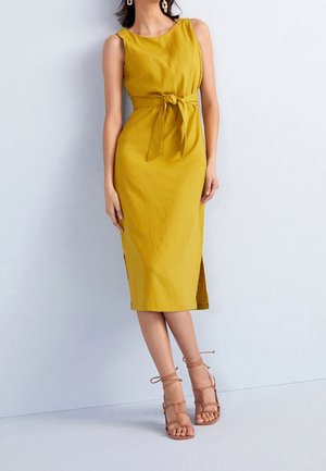 Femme portant une robe midi jaune sans manches avec un nœud de ceinture et des sandales beige à talons hauts lacées devant un mur uni.